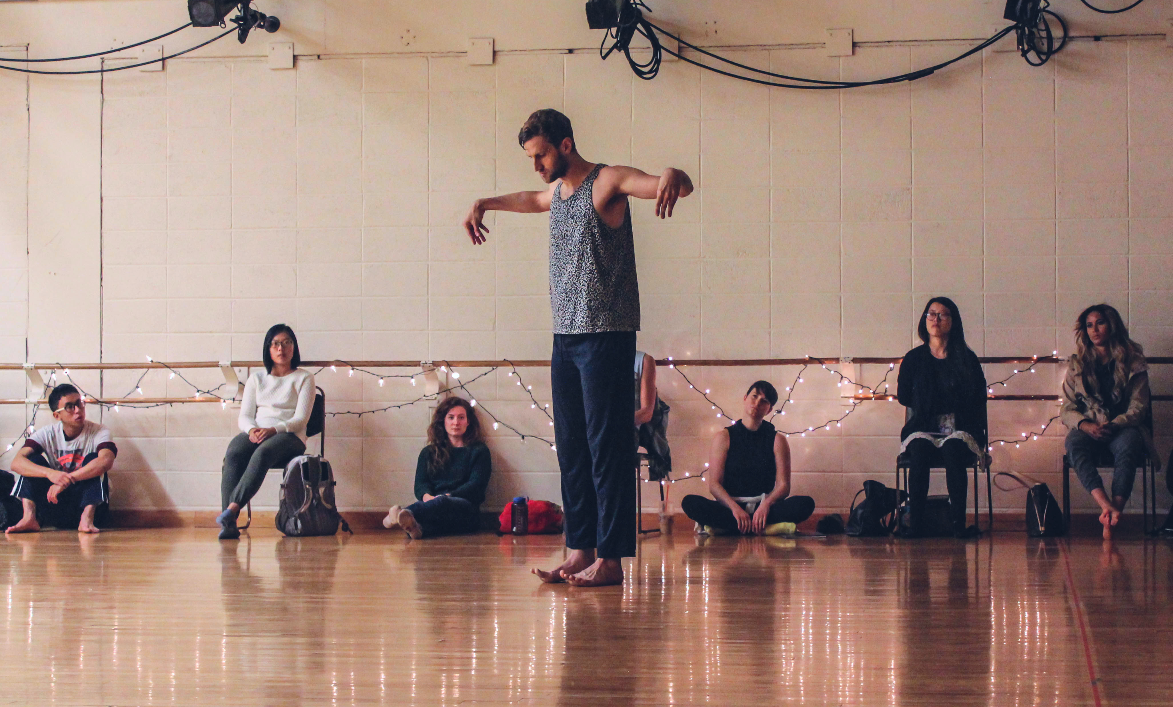 Dancer Brendan P. Behan, a tall, white cisgender male, stands upright performing in a low-lit dance studio on a wooden floor. His arms are held out to the side, elbows bents, forearms extending forward from the elbow at a ninety-degree angle, hands cupped palms-down, and his back is angled toward to the audience behind him at a roughly 45-degree angle. His head is tilted forward as he looks down at the ground where his bare feet, both in contact with the floor, curl upward from the toes lifting off the floor below. Behind him are audience members variously sitting on the floor and in chairs in front of a brick wall. The wall has a double wooden ballet barre affixed to it with white string lights, which are lit, woven through it.