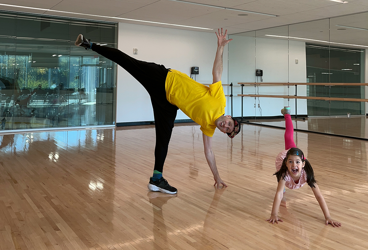 A dance student smiles at the camera while demonstrating a penché style leg lift pose while their teacher performs a sideways tilt balancing on one leg inside the dance studio at the Robert Crown Community Center.