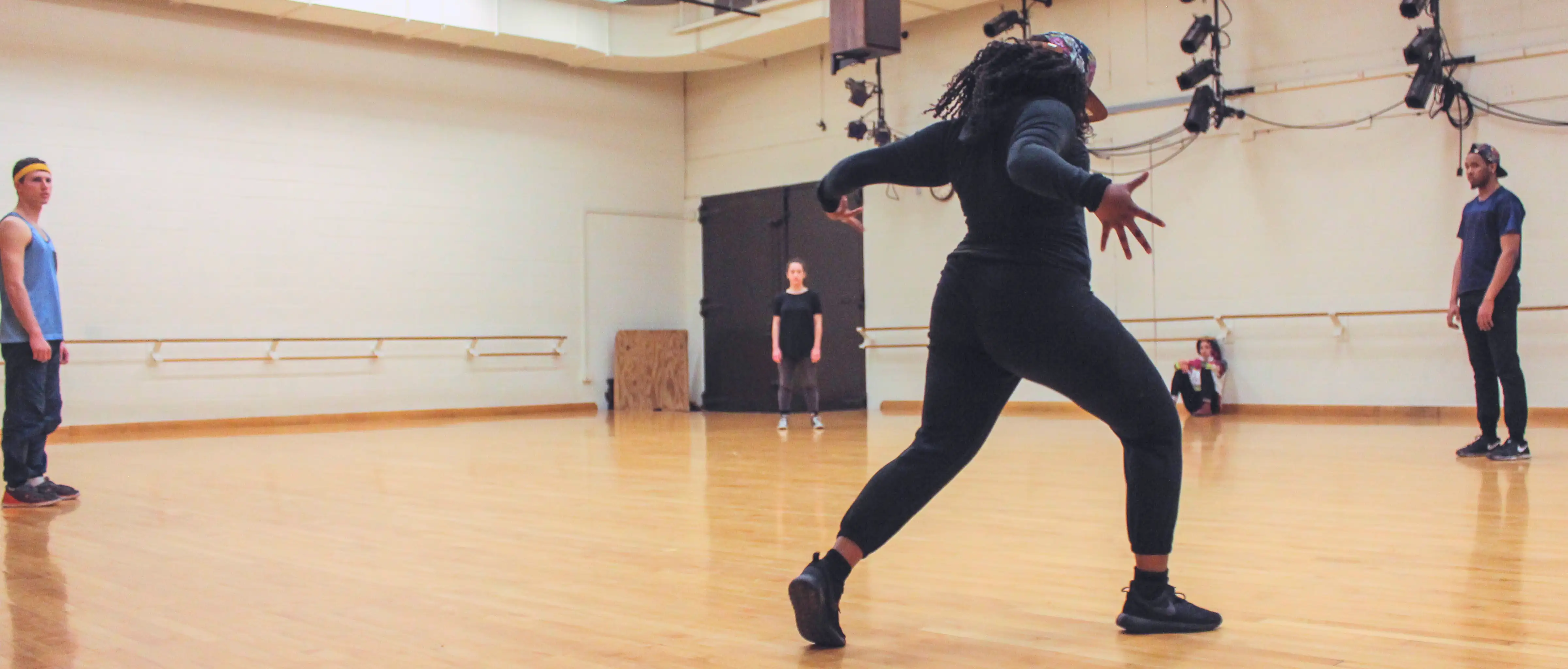 Four dancers standing far apart in a wide circle watch as the dancer in the foreground moves toward the center of the floor as she performs an emphatic, dynamic krump movement with arms wide and feet apart as part of Behan's dance work 'Pxyxl OR ...'. A Fifth dancer is seating and watching off to the far right side.