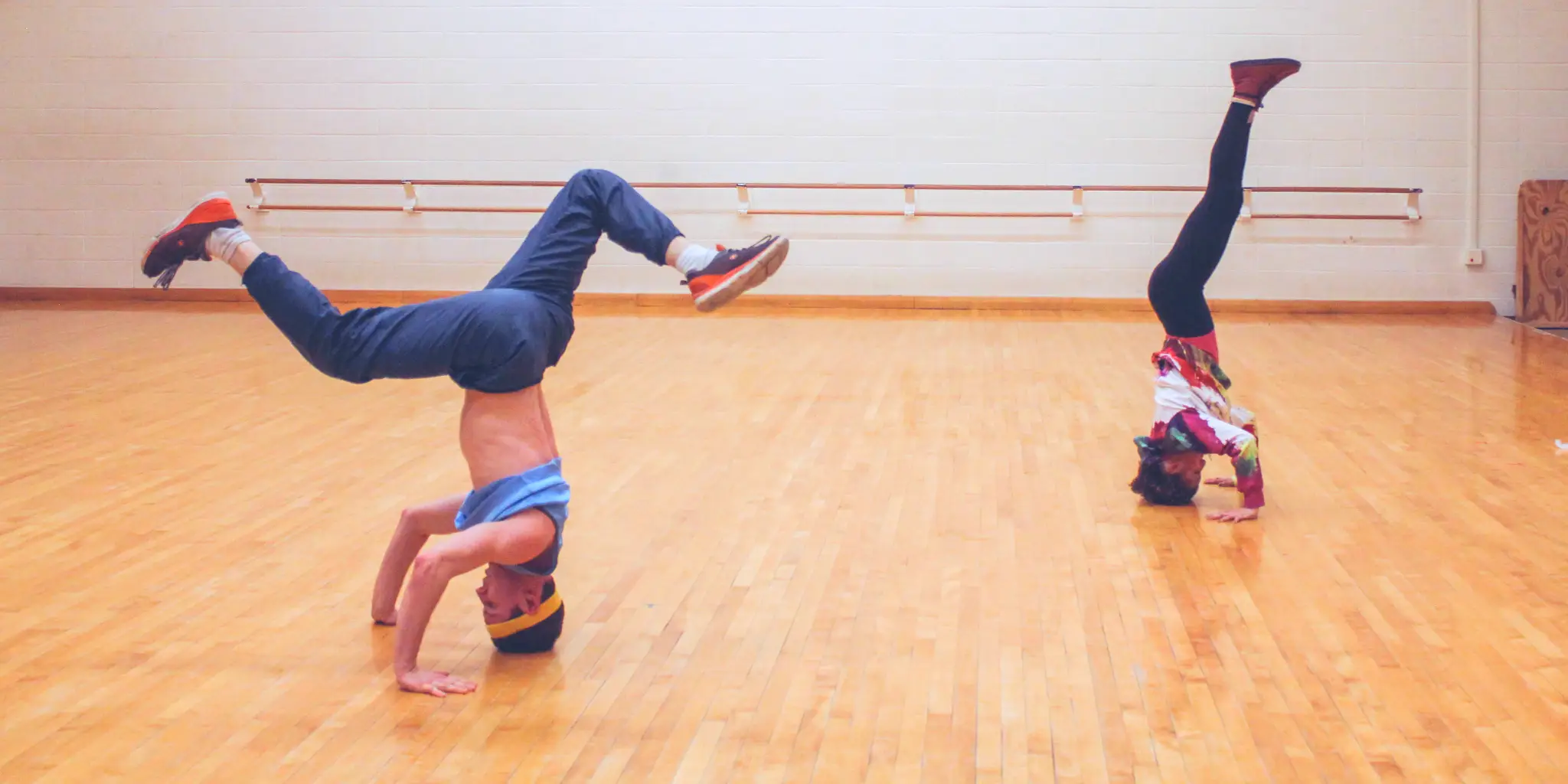 A photo of two dancers, one male presenting and the other female-presenting, spaced apart, both on the floor, heads to the ground doing various kinds of breakdance balances with their legs in the air as they perform in a dress rehearsal for Behan's work 'Pxyxl OR Padrao e Compasso OR ...'.
