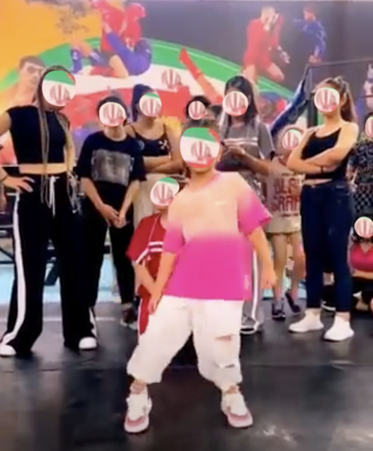 A young girl dancer in the foreground wearing western baggy clothing gets ready to dance in front of a group of other youth in a room that looks like a recreational center.