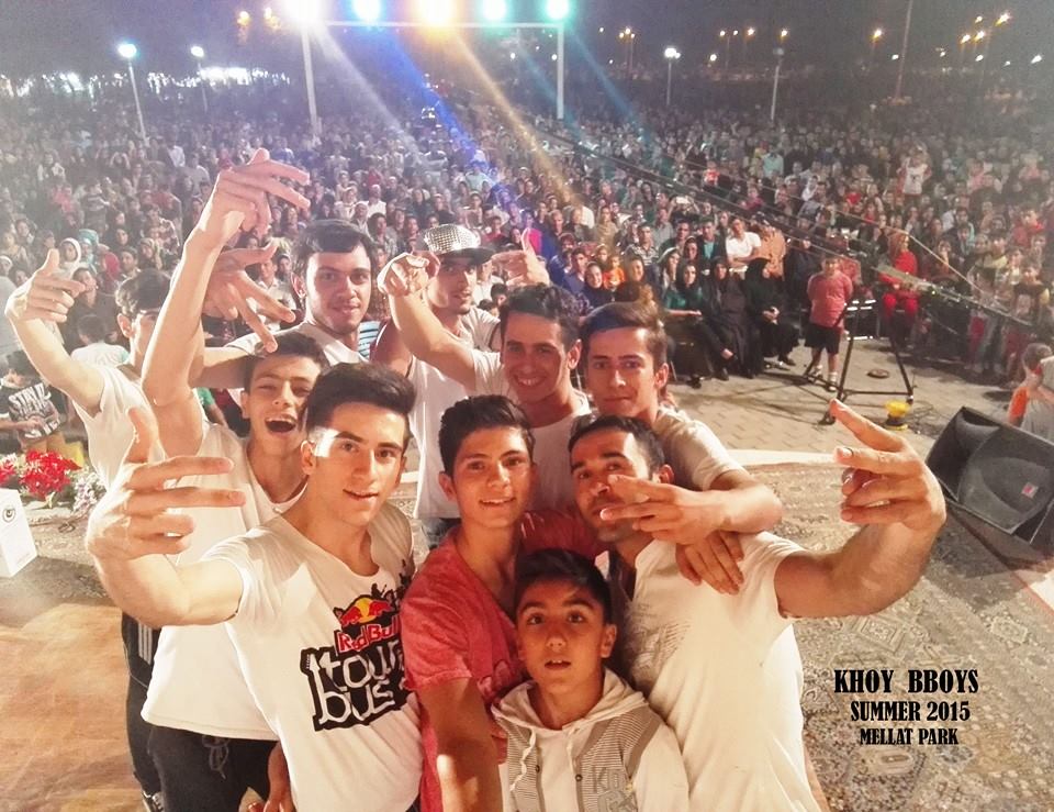 Khoy B-Boys grouped close together in the foreground pose arms up and smiling on a dance floor in front of a large crowd in Mellat Park in Tehran