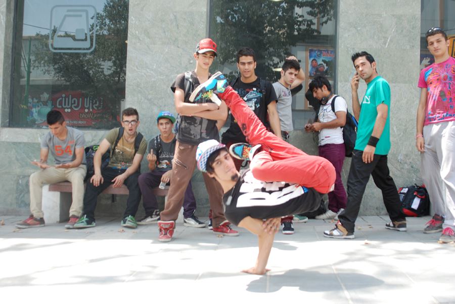 A male hip hop dancer wearing Western-style clothing and a baseball hat demonstrates a breaking freeze in which he balances on a single arm which is bent under his back while his torso faces upward and he kicks one leg up into the air with the other leg bent, turned outward, and hooked around the straight leg. The dances is on a sidewalk in Tehran on a sunny day and a group of boys variously standing and sitting outside of a building window look on toward the dancer in the foreground.