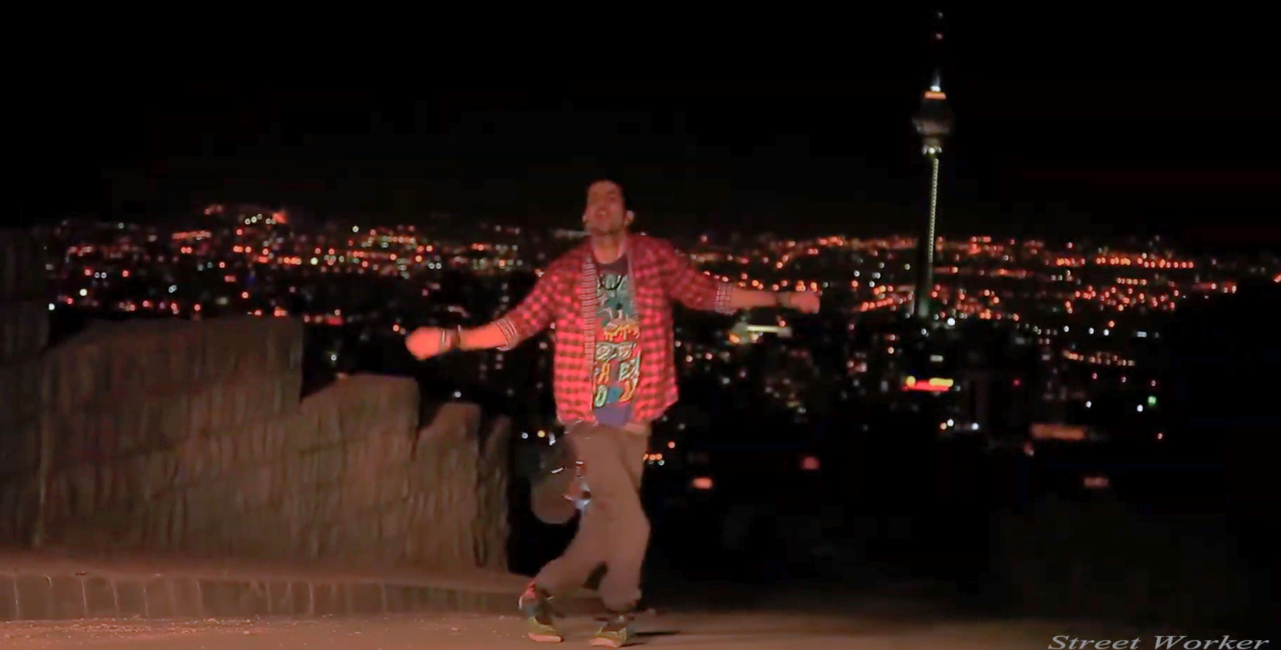 A male-presenting Iranian dancer dancing in a fusion hip hop style standing upright, legs crossed, knees bent, and arms out, head titled back, on a street at night with the Tehran cityscape visible in the background including the lights of Milad Tower