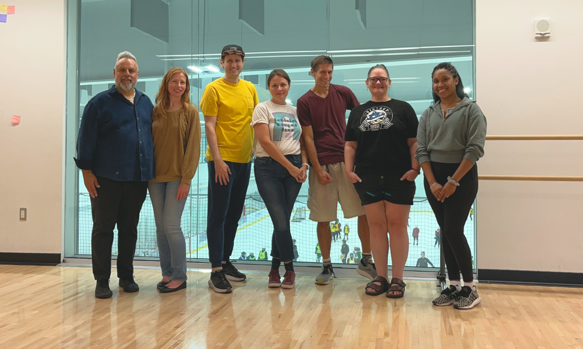 A class photo with adult students and the teacher standing choulder to shoulder in line in front of a ceiling-to-floor window which overlooks an ice rink down below where a hockey game is in progress.