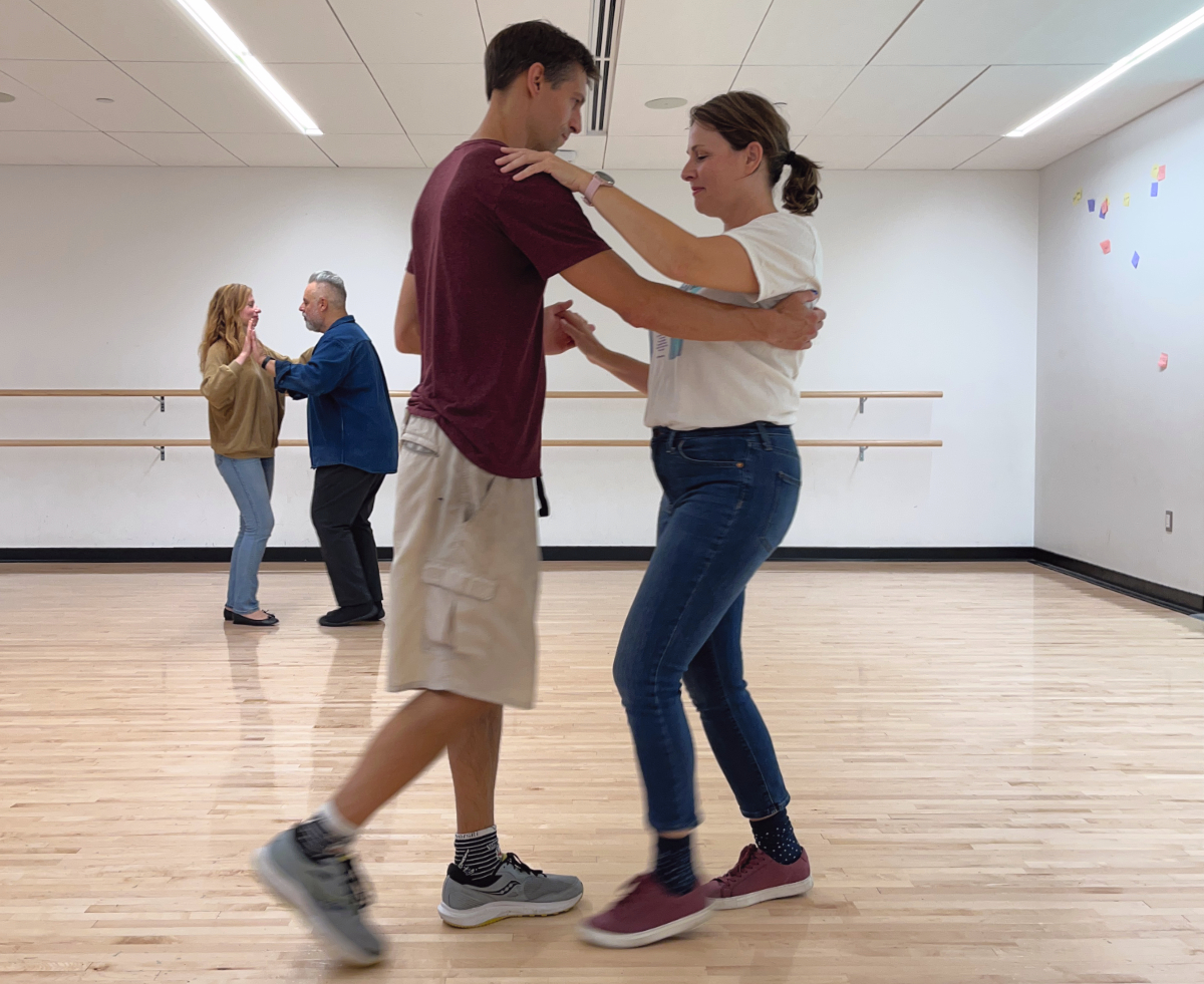 Two couples, one in the foreground and one in the background, dance with each other, arms intertwined on one side, hands connected on the other, and facing each other, inside the dance studio at the Robert Crown Community Center.