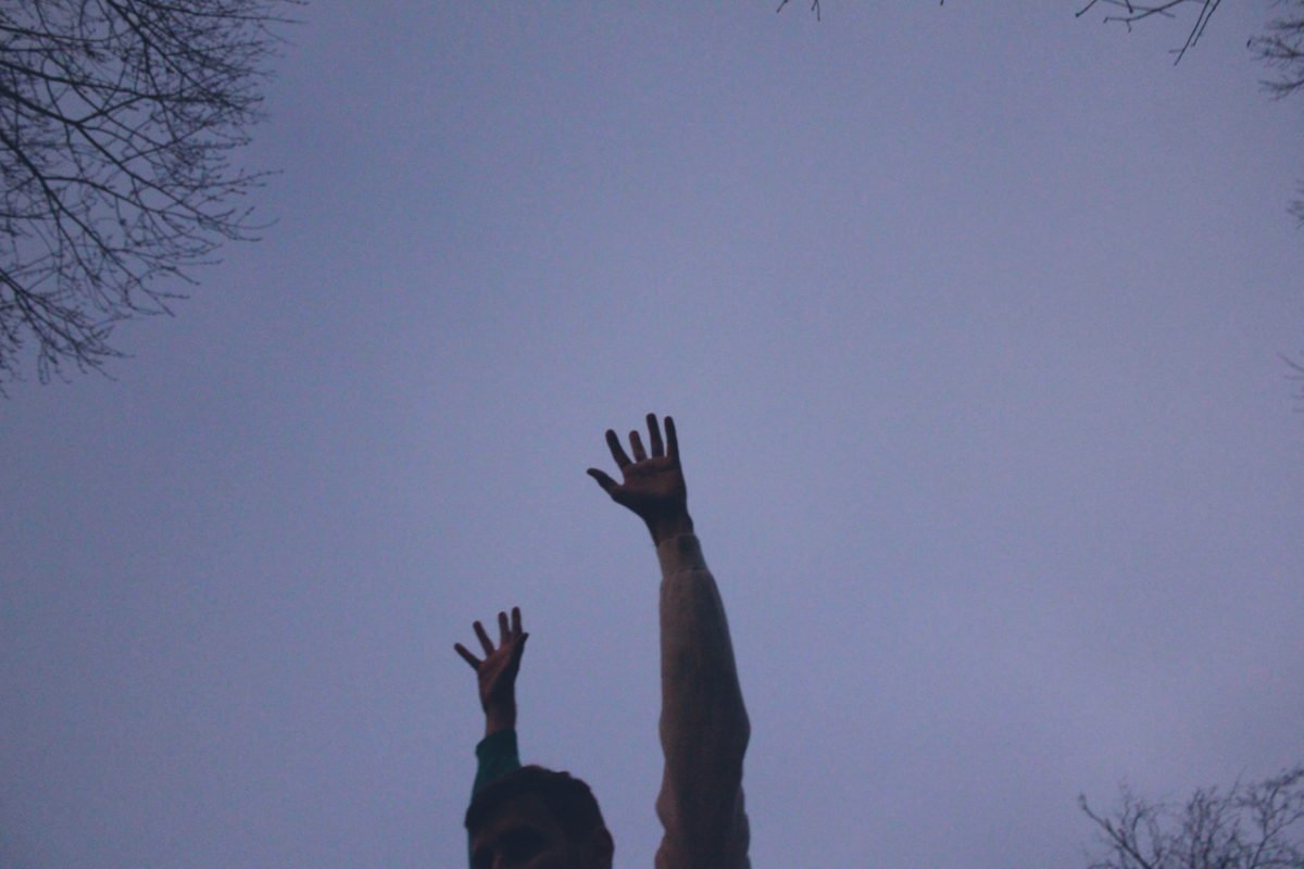 A photo. Silhouettes of three leafless trees appear in the corners against a darkened gray-blue dusk sky. In the foreground, dancer Brendan P. Behan raises both arms skyward, hands open and fingers apart, as he looks out of the image frame to the viewer's left. His image is cut off by the bottom of the image frame and shows only his arms and half of his face, which are not well lit and dark to the point of detail and color being difficult to make out. Brendan is wearing a sweater which covers his arms.