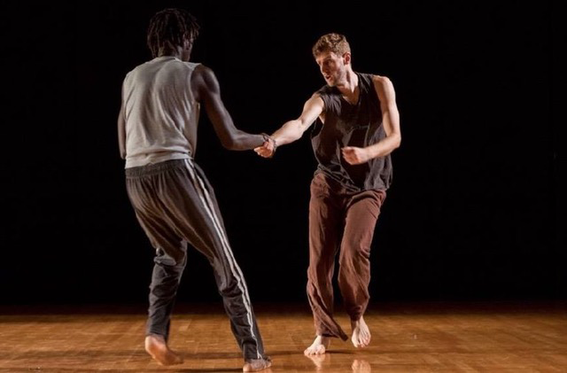 Dancers Momar Ndiaye and Brendan P. Behan, a tall Black man and a tall white man, respectively, performing on stage, grasping each other with their right arms and facing each other with an intense gaze as they lean away from each other.